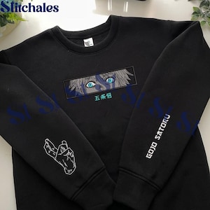 Op de afbeelding: Zwart sweatshirt met borduursel in anime-stijl. Het ontwerp toont de ogen en het haar van een personage, met Japanse tekst eronder. De linkermouw heeft een handgebaar en de rechtermouw zegt "GOJO SATORU" in het wit.