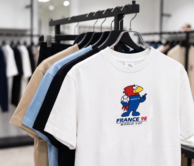 May include: A white short-sleeved t-shirt featuring a colorful graphic of a cartoon rooster holding a soccer ball with the text FRANCE 98 WORLD CUP. The shirt is displayed on a black hanger alongside other t-shirts in tan, blue, and black.