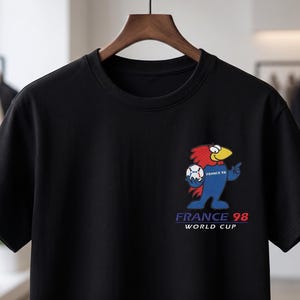 May include: A black short-sleeve t-shirt featuring a graphic of a cartoon rooster holding a soccer ball. The design includes the text "FRANCE 98" and "WORLD CUP" in blue and red lettering, positioned on the left chest area of the garment.
