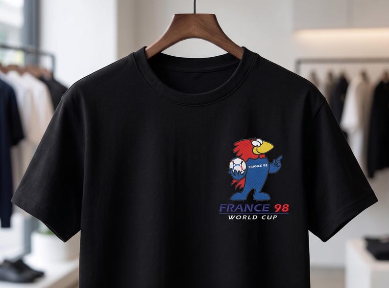 May include: A black short-sleeve t-shirt featuring a graphic of a cartoon rooster holding a soccer ball. The design includes the text "FRANCE 98" and "WORLD CUP" in blue and red lettering, positioned on the left chest area of the garment.