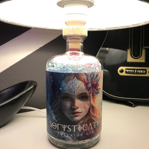 Può includere: Una bottiglia di gin in vetro trasparente con tappo in sughero, con un'etichetta con un ritratto di donna e la scritta "SØGYSTØCATE SUPERIOR GIN". La bottiglia è sotto una lampada.