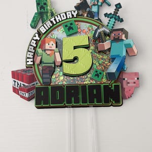 May include: A Minecraft-themed birthday cake topper with the text "Happy Birthday" and the number "5". The topper features Minecraft characters, including Steve, Alex, a Creeper, and a pig. The name "Adrian" is also displayed.