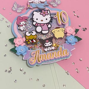 Puede incluir: Un adorno para pastel de capas rosa, azul y morado con un personaje de Hello Kitty, una rana, un perro y un gato blanco y negro. El adorno también incluye un avión, mariposas y el nombre "Amanda" en letras doradas. El número "10" también está incluido en letras doradas.