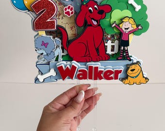 May include: A colorful birthday cake topper featuring Clifford the Big Red Dog, balloons, and the number 2. The topper includes the name "Walker" and other cartoon characters. The design is held by clear plastic sticks.