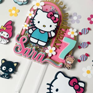 Puede incluir: Un adorno para pastel rosa y dorado con un personaje de Hello Kitty, el número 7 y las palabras "Feliz cumpleaños Sara".