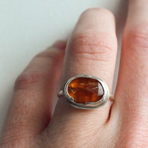 May include: A silver ring featuring a large, faceted, amber-colored gemstone. The stone is oval-shaped and set in a simple bezel setting. The ring is shown on a finger against a white background.