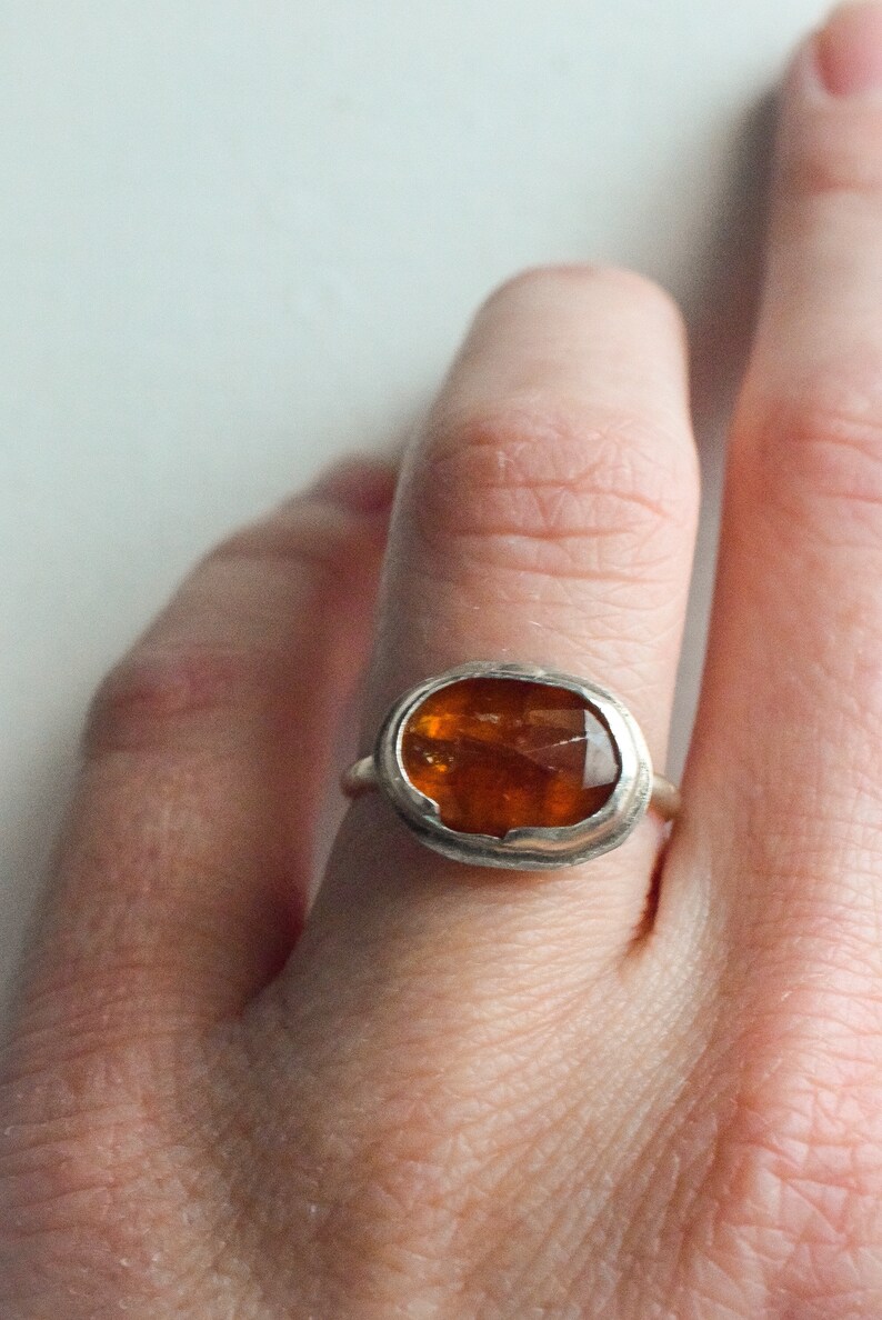 May include: A silver ring featuring a large, faceted, amber-colored gemstone. The stone is oval-shaped and set in a simple bezel setting. The ring is shown on a finger against a white background.