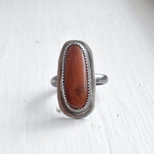 May include: A silver ring featuring a large, oval, reddish-brown stone. The stone is set within a serrated bezel, and the ring has a simple band. The ring is displayed on a white surface.