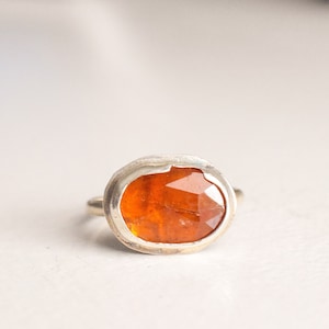 May include: A silver ring featuring a faceted, oval-shaped orange gemstone. The gemstone is set in a bezel setting, and the ring has a simple, polished band. The ring is displayed on a white surface.