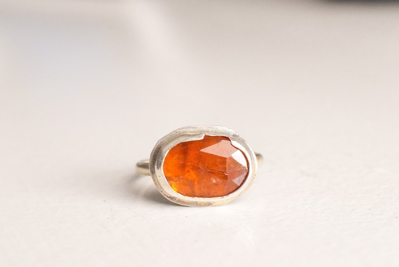 May include: A silver ring featuring a faceted, oval-shaped orange gemstone. The gemstone is set in a bezel setting, and the ring has a simple, polished band. The ring is displayed on a white surface.
