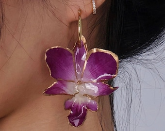 May include: A pair of gold hoop earrings with a purple and white orchid flower design. The earrings are made of metal and have a gold finish.