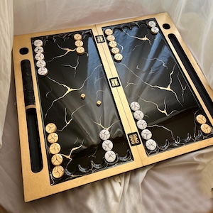 Może przedstawiać: Czarno-złota plansza do gry w backgammona z białymi i złotymi pionkami. Plansza wykonana jest z drewna i ma marmurowy wzór.