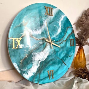 Puede incluir: Un reloj redondo grande con un diseño abstracto turquesa y blanco. La esfera del reloj tiene números romanos y manecillas doradas.