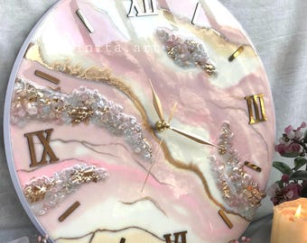 May include: Round wall clock with a white frame, featuring a pink, white, and gold abstract design. Roman numerals mark the hours. The clock hands are gold. The clock has a resin finish and decorative elements.