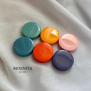 May include: Six smooth, circular resin cabochons in teal, orange, yellow, pink, dark blue, and red-orange. The objects are arranged in a cluster. The text RESINITA Est. 2020 is printed in black in the lower left corner of the image area.