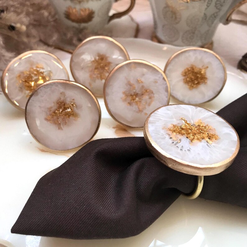 May include: A collection of white and gold napkin rings. Each ring features a circular design with a white center and gold flecks, encased in a gold-colored metal rim. One ring is holding a dark brown napkin.