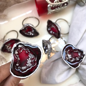 May include: A collection of decorative napkin rings. Each ring features a dark red, geode-like design with silver accents and a silver ring. The napkin rings are arranged on a white surface, with a white napkin in the background.