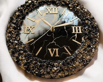 May include: A black and gold resin clock with Roman numeral markings. The clock face has a white and black marble design with gold accents.