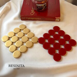 May include: Two square arrangements of circular coasters, one set in a metallic gold finish and the other in a glossy red finish. The text RESINITA Est. 2020 is printed in black in the lower left corner, with a red book and candle above.