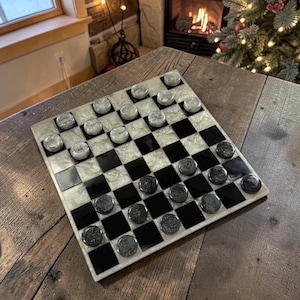 May include: A checkerboard game with black and white squares and clear and black checkers. The board has a pearlescent, marbled effect. The game is set on a rustic wooden table, with a fireplace and Christmas tree in the background.