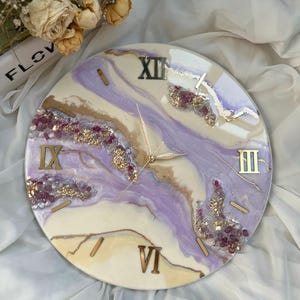 Pode incluir: Um relógio redondo com fundo branco e design abstrato roxo e dourado. O mostrador do relógio tem algarismos romanos e ponteiros dourados.