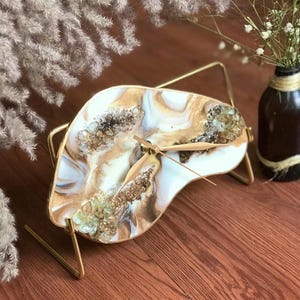 May include: A white and gold resin clock with gold glitter and clear quartz crystals embedded in the resin. The clock is on a gold metal stand.