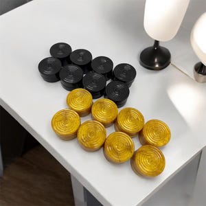 Op de afbeelding: Een set damstenen met zwarte en gouden stukken. De damstenen zijn in twee rijen op een wit oppervlak gerangschikt.