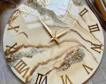 May include: A white and gold resin clock with Roman numeral markings. The clock face has a marbled design with gold accents and clear crystals.