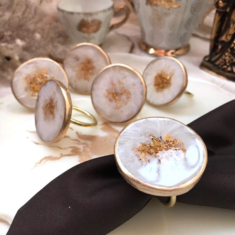 May include: A set of six decorative napkin rings. Each ring features a white and gold design with gold flakes. The rings are round with a gold-colored metal band. One ring is placed on a black napkin.