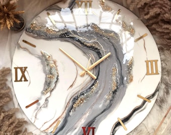 May include: A large, round, white clock with Roman numeral markings and gold accents. The clock face features a gray and white abstract design with gold glitter accents.