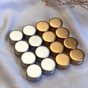 May include: An assortment of small, round metal tins in silver and gold colors. The tins are arranged in a neat pattern on a white, textured surface. The lids are flat and the sides are cylindrical.