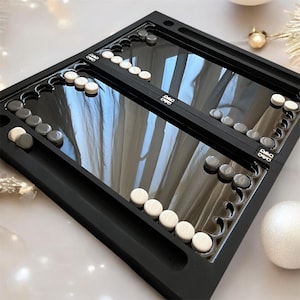May include: A black wooden backgammon board with a mirrored playing surface. The board is open and shows white and grey playing pieces.