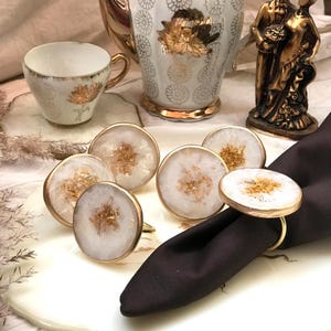 May include: A collection of white and gold agate napkin rings with gold rims and accents. The rings are arranged on a white plate with a black napkin. A teacup, teapot, and decorative figurine are in the background.