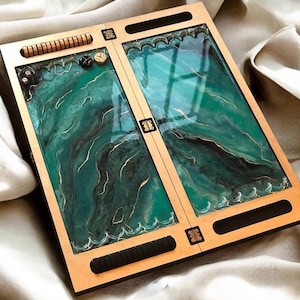 May include: A backgammon board with a teal and black marbled design. The board is framed in light wood with black accents and a decorative border. Dice and game pieces are visible. The rectangular board has a glossy finish. The board measures approximately 40 cm x 30 cm.