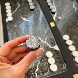 May include: A black and silver game piece with a glittery surface. The piece is being held by a hand.