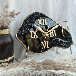 Könnte beinhalten: Schwarze und goldene Harz-Uhr mit römischen Ziffern auf einem goldenen Metallständer. Das Zifferblatt der Uhr ist unregelmäßig geformt und hat einen Marmoreffekt.