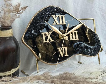 May include: Black and gold resin clock with Roman numeral markings on a gold metal stand. The clock face is irregularly shaped and has a marbled effect.