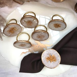 May include: A set of six decorative napkin rings. Each ring features a gold-colored metal loop attached to a white and gold resin oval. The resin has a geode-like appearance with gold flecks. The napkin rings are arranged on a white and gold decorative tray.