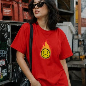 May include: A red t-shirt featuring a graphic of a yellow, sad-faced emoji engulfed in flames. The shirt is worn with blue jeans and a black backpack. The background includes red crates and urban graffiti.