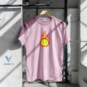 May include: A light pink t-shirt with a graphic of a yellow, sad-faced emoji engulfed in red and orange flames. The letters "C G" are inside the emoji. The shirt is hanging on a wooden hanger.