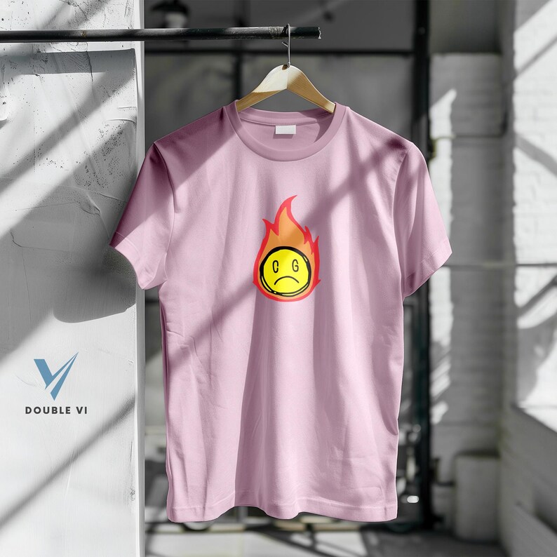 May include: A light pink t-shirt with a graphic of a yellow, sad-faced emoji engulfed in red and orange flames. The letters "C G" are inside the emoji. The shirt is hanging on a wooden hanger.