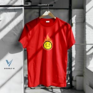 May include: A red t-shirt with a graphic of a yellow, sad-faced emoji surrounded by orange flames. The letters "C G" are on the emoji. The shirt is hanging on a wooden hanger.