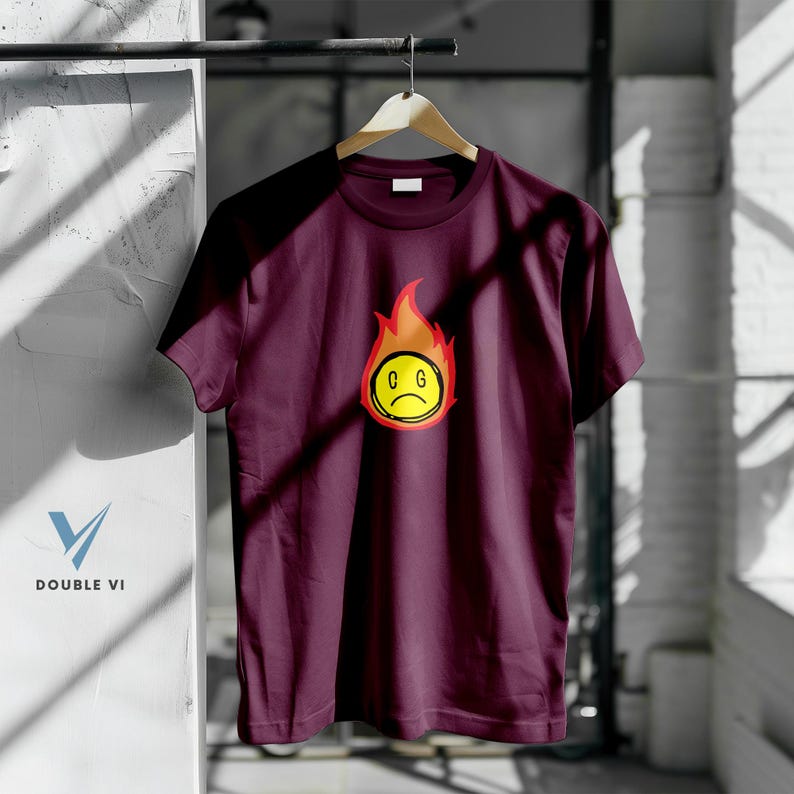 May include: A maroon t-shirt with a graphic of a yellow, sad-faced emoji engulfed in orange and red flames. The emoji has the letters "C G" inside the yellow circle. The shirt is hanging on a wooden hanger.