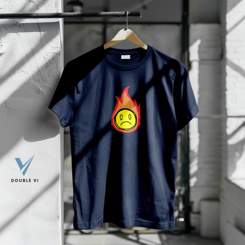 May include: Navy blue t-shirt with a graphic of a sad face inside a flame. The face is yellow with a red outline and the letters "C G" are below the face. The flames are red and orange. The shirt is hanging on a wooden hanger.