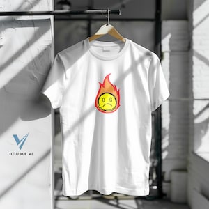 May include: White t-shirt with a graphic of a yellow, sad-faced emoji surrounded by orange and red flames. The letters "C G" are on the emoji. The shirt is hanging on a wooden hanger.