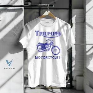 May include: White t-shirt featuring a blue graphic of a Triumph motorcycle. The word "TRIUMPH" is printed above the motorcycle, and "MOTORCYCLES" is printed below. The shirt is hanging on a wooden hanger.
