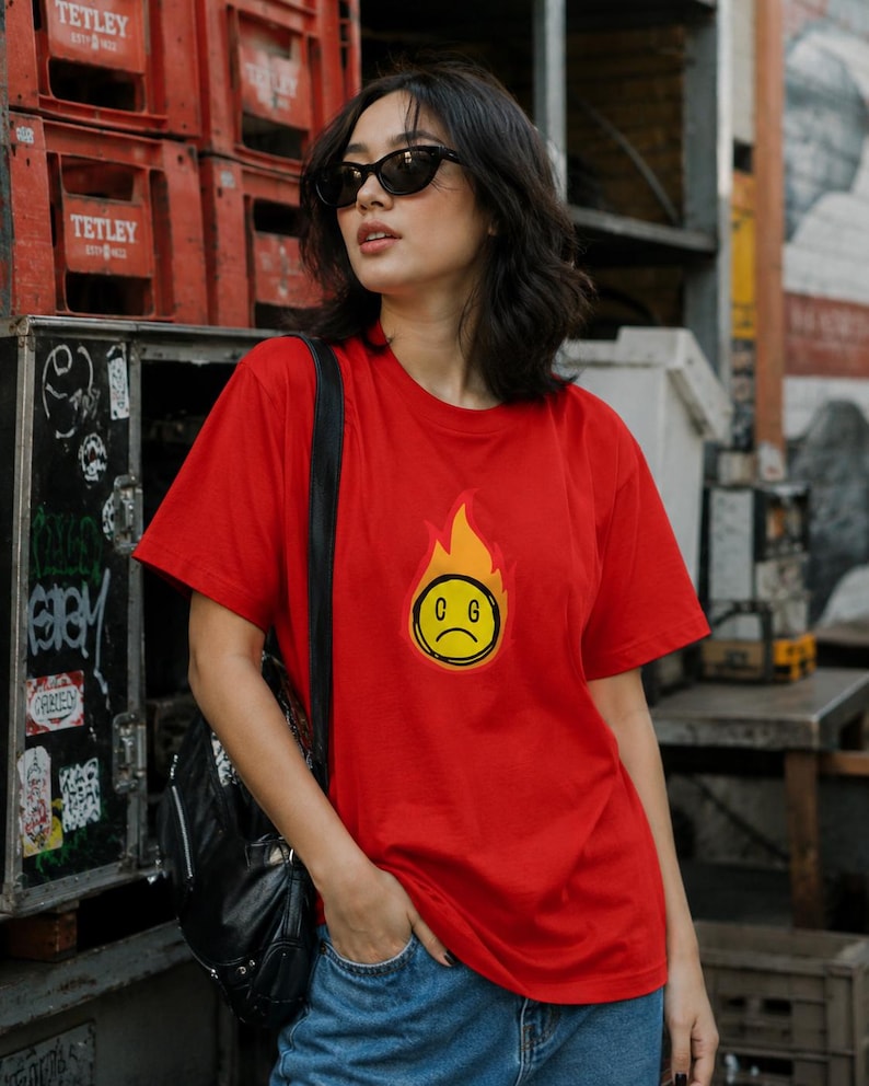 May include: A red t-shirt featuring a graphic of a yellow, sad-faced emoji engulfed in flames. The shirt is worn with blue jeans and a black backpack. The background includes red crates and urban graffiti.