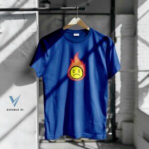 May include: A blue t-shirt with a graphic of a yellow, sad-faced emoji surrounded by orange and red flames. The emoji has the letters "C G" inside the yellow circle. The shirt is hanging on a wooden hanger.