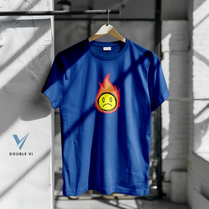 May include: A blue t-shirt with a graphic of a yellow, sad-faced emoji surrounded by orange and red flames. The emoji has the letters "C G" inside the yellow circle. The shirt is hanging on a wooden hanger.