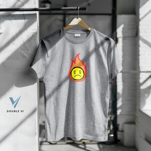 May include: A heather gray t-shirt with a graphic of a sad yellow face engulfed in red and orange flames. The letters "C G" are inside the yellow face. The shirt is hanging on a wooden hanger.